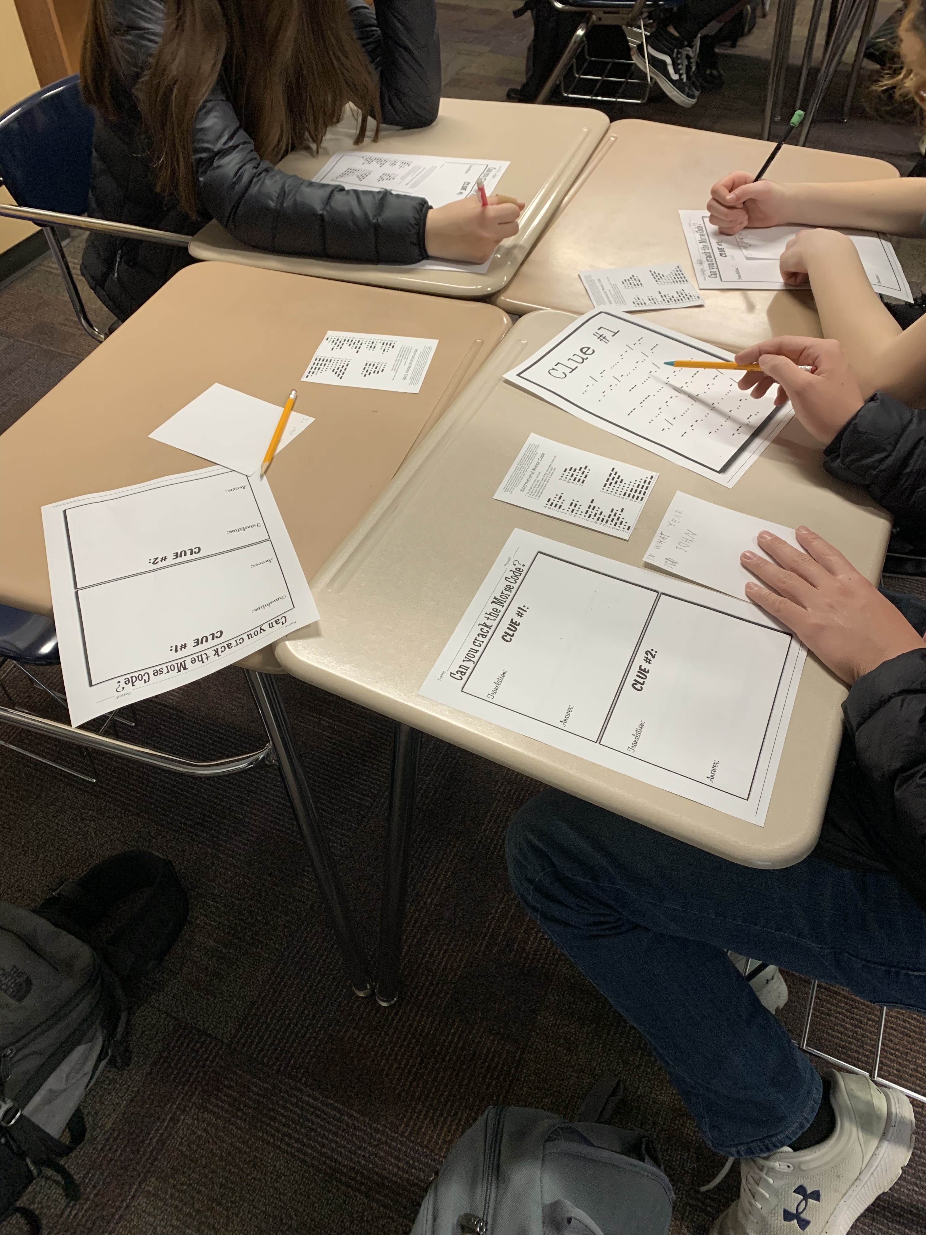 Four students seated at desks working on worksheets labeled 'Clue 1' and 'Clue 2,' with small printed paper slips and pencils on the desks